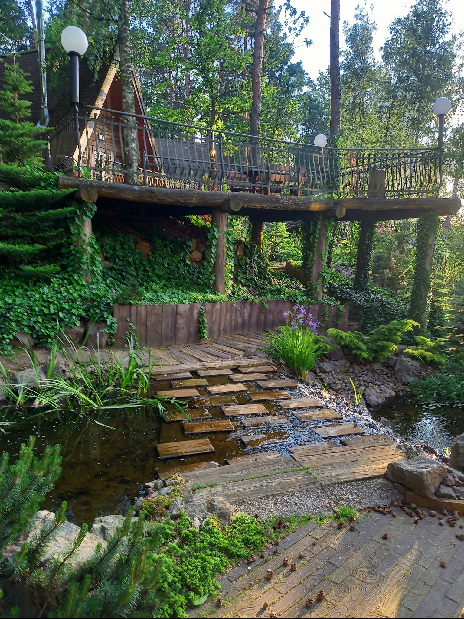 Ogród z drewnianym pomostem nad strumieniem, prowadzącym do podwyższonej platformy z balustradą i domkiem na drzewie w tle, otoczony bujną zielenią i lampami ogrodowymi.