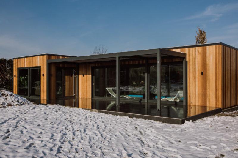 Nowoczesna, drewniana dobudówka z dużymi oknami i zadaszeniem tarasowym, otoczona śniegiem.