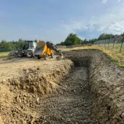 Wąski wykop w ziemi z gruzem, wykonany równolegle do ogrodzenia z siatki, z widocznymi maszynami budowlanymi w tle i zielenią drzew na horyzoncie pod błękitnym niebem.
