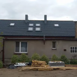 Dom z nowym, grafitowym dachem z blachy trapezowej, widoczne okna dachowe i dwa kominy, na ziemi leżą nowe dachówki i drewno, drabina oparta o ścianę.