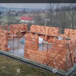 Wznoszone ściany z czerwonej cegły na fundamencie, widok na częściowo wybudowany dom w surowym stanie, na tle krajobrazu z drzewami i zabudowaniami.