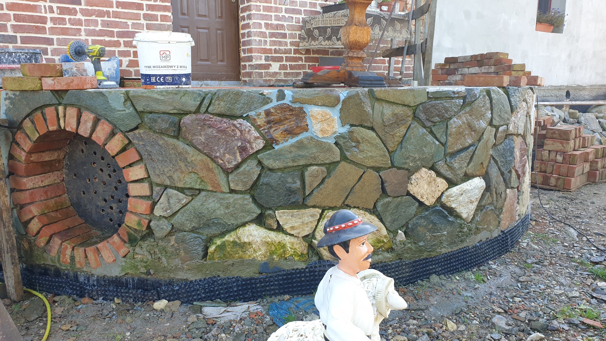 Obróbka kamienia elewacyjnego i cegły klinkierowej na fundamencie domu z otworem wentylacyjnym, widoczna figura dekoracyjna w stroju regionalnym.