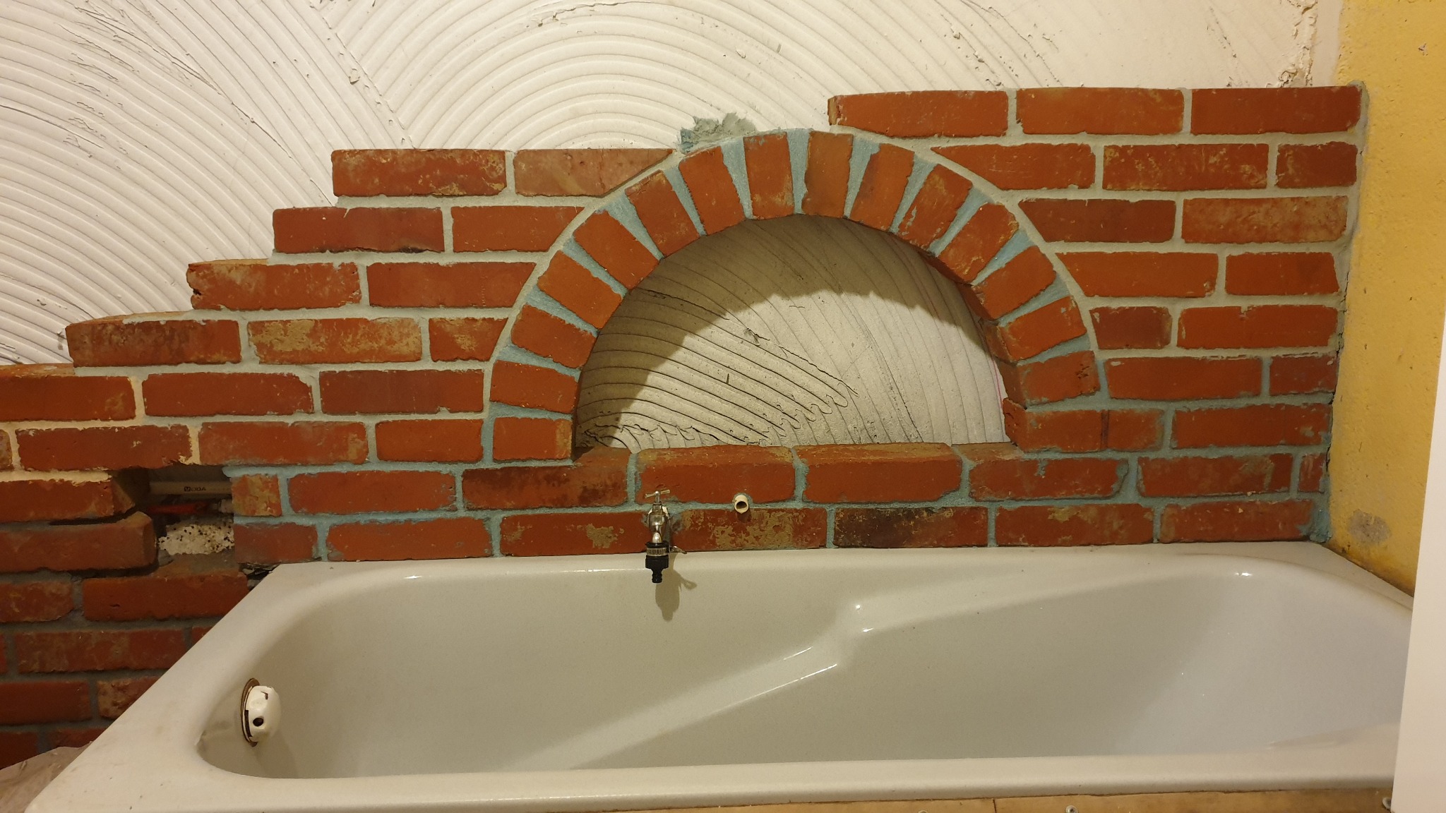 Niedokończona ściana z czerwonej cegły nad wanną z zaokrąglonym otworem, widoczne ślady zaprawy na ścianie i cegłach.