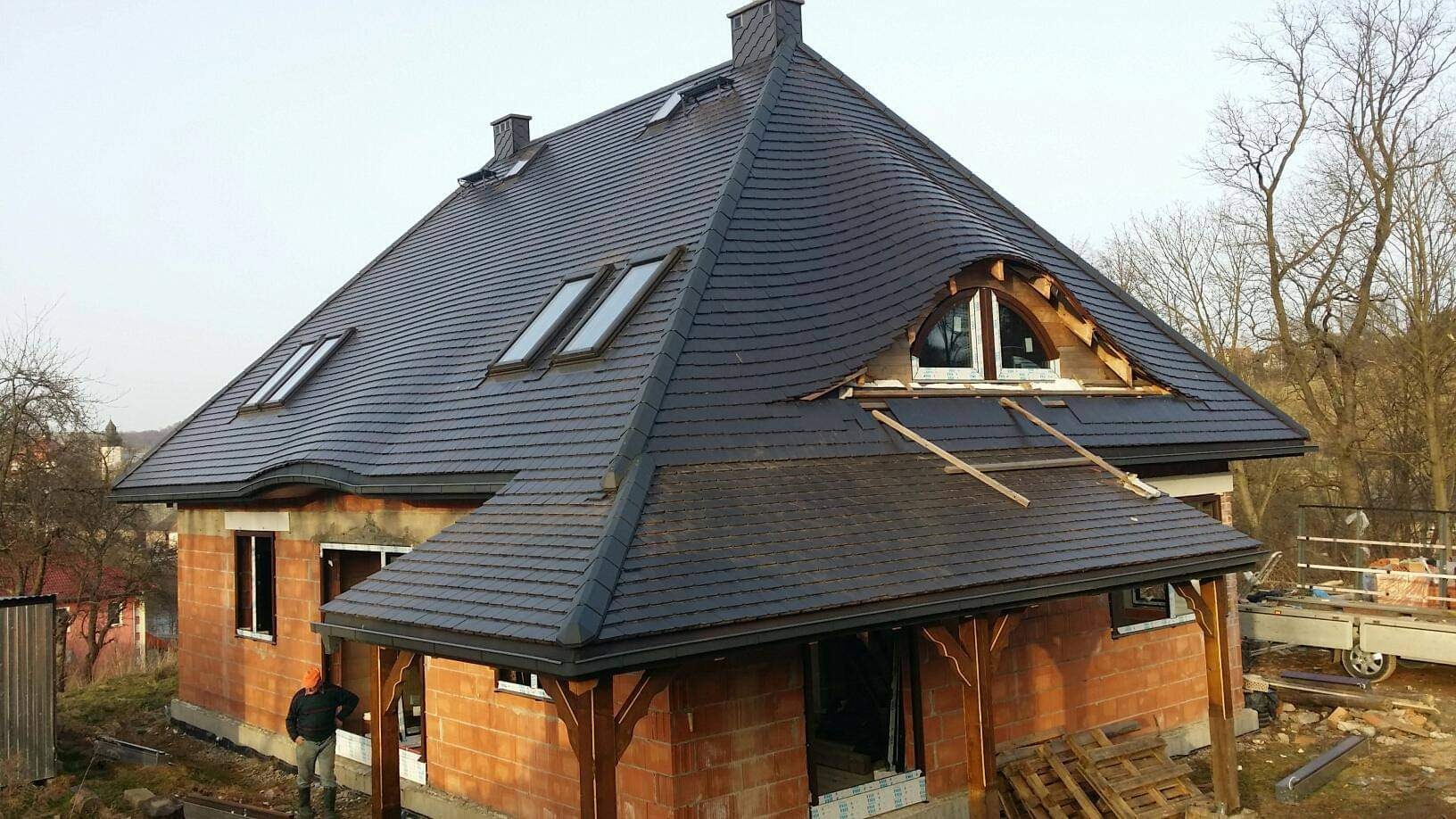 Niedokończony dom z ciemną, łuskową dachówką, widoczne okna dachowe i okrągłe okno w szczycie, ceglana elewacja w trakcie budowy, pracownik w kasku i roboczych butach.