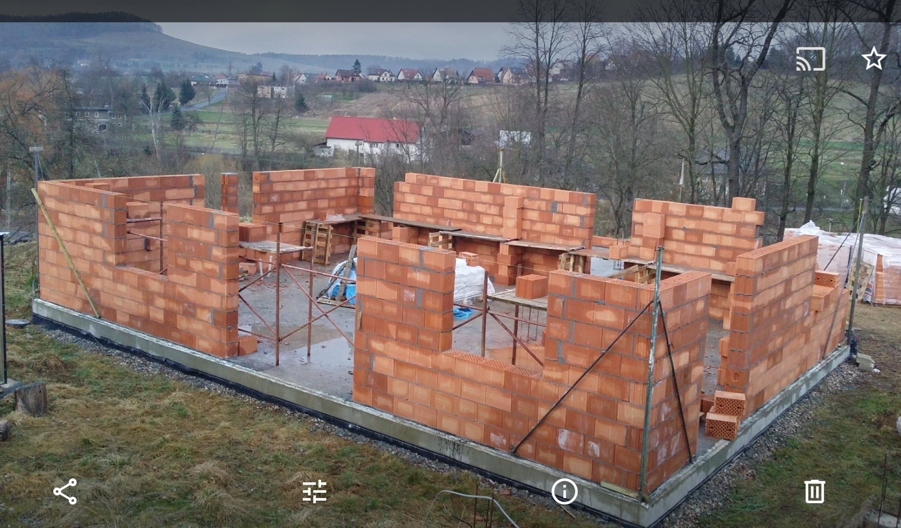Wznoszone ściany z czerwonej cegły na fundamencie, widok na częściowo wybudowany dom w surowym stanie, na tle krajobrazu z drzewami i zabudowaniami.