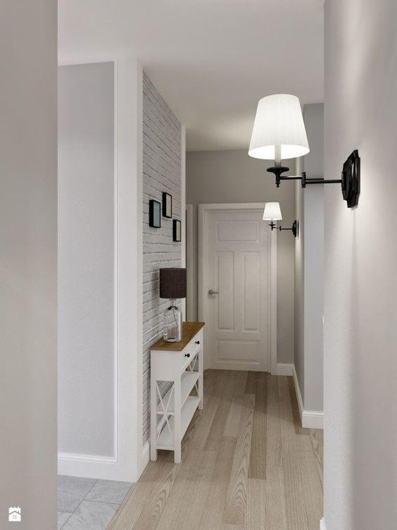 Jasne wnętrze przedpokoju z drewnianą podłogą, białą konsolą z lampką, ceglaną ścianą i czarnymi kinkietami z abażurami.
