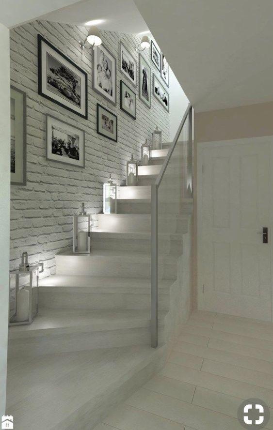 Schody z białego drewna z szklaną balustradą, oświetlone lampionami, ściana z cegły pomalowana na biało, udekorowana czarno-białymi fotografiami w ramkach.