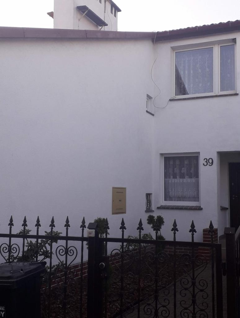 Biała elewacja domu jednorodzinnego z numerem 39, widoczna skrzynka pocztowa i fragment ozdobnego, czarnego ogrodzenia z przodu.
