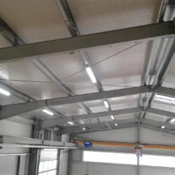Wnętrze hali przemysłowej z widoczną konstrukcją stalową dachu, oświetleniem jarzeniowym i suwnicą pod stropem.