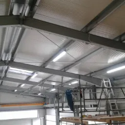 Wnętrze hali przemysłowej z widoczną konstrukcją stalową dachu, oświetleniem LED i elementami instalacji elektrycznej w trakcie montażu, widoczne rusztowanie i drabina.