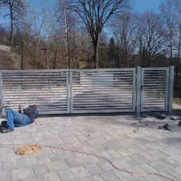 Spawanie metalowej bramy przesuwnej z poziomymi szczeblami na posesji z kostki brukowej; spawacz w niebieskich jeansach leży na ziemi, a obok leżą narzędzia i zwinięty kabel.