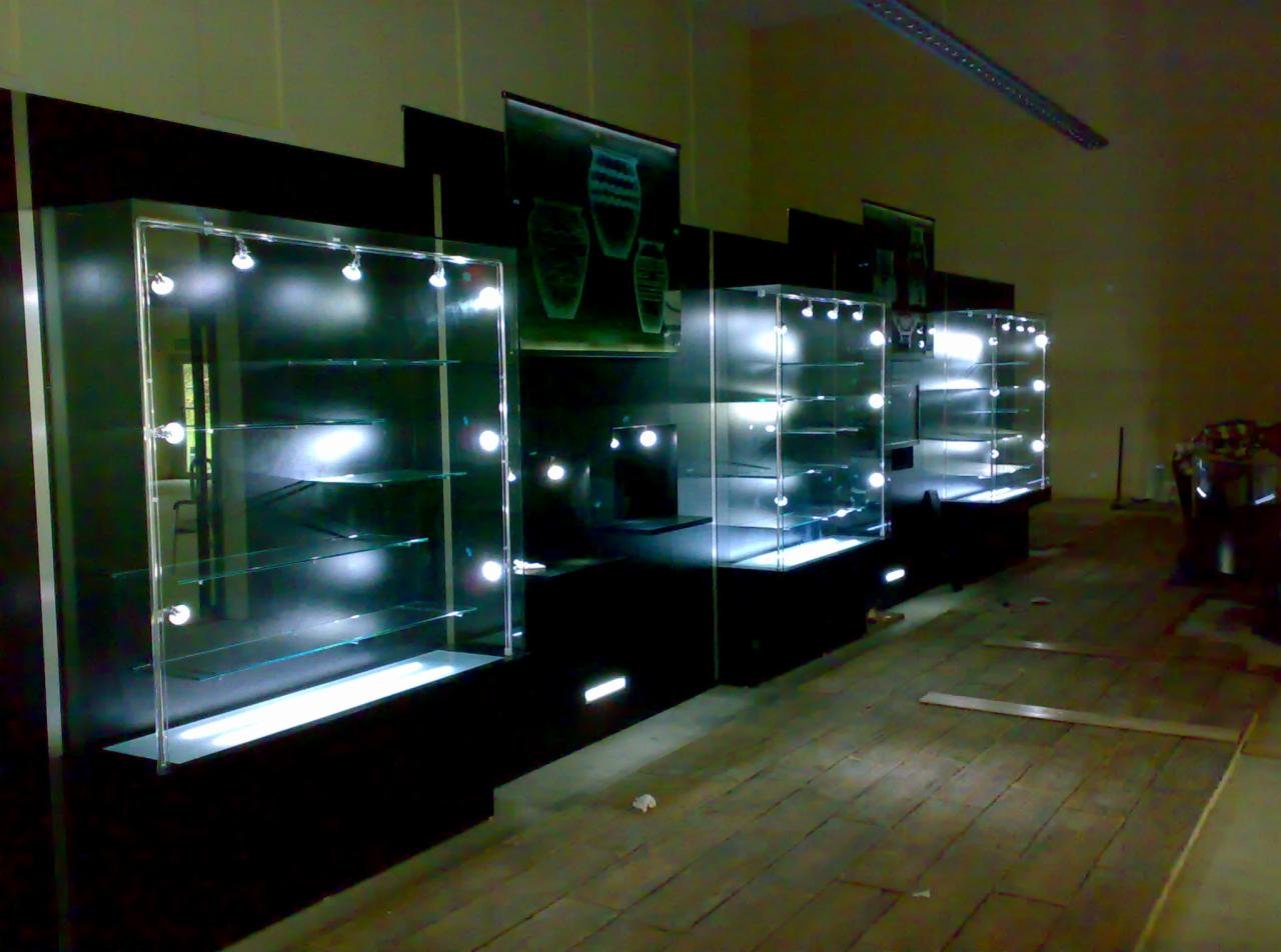 Rząd podświetlanych, szklanych gablot ekspozycyjnych z punktowym oświetleniem LED, zamontowanych na czarnych postumentach, z grafiką na ścianie w tle, w trakcie instalacji, widoczne elementy...