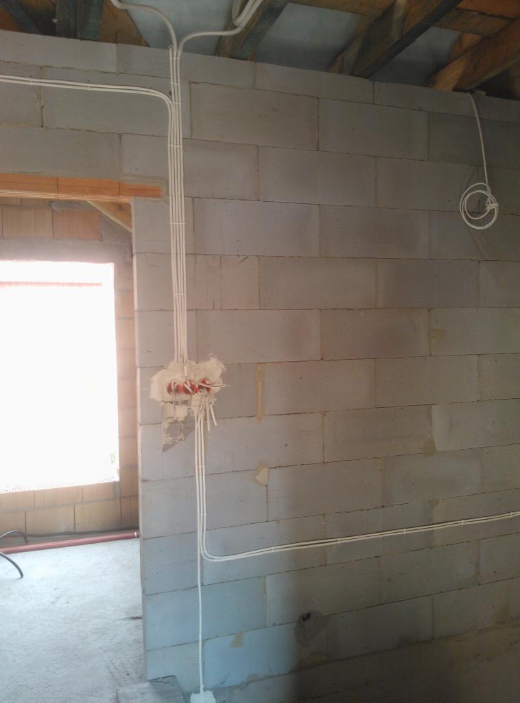 Surowe wnętrze w trakcie prac: białe przewody prowadzone po ścianie z bloczków betonowych, widoczne puszki z przewodami, fragment okna w tle.