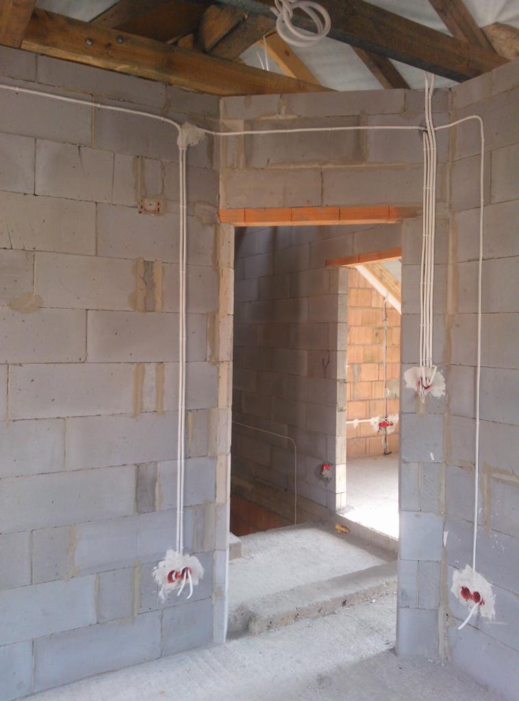 Wewnętrzne ściany z betonu komórkowego w trakcie prac: białe przewody poprowadzone po ścianach, przygotowane punkty pod gniazdka i włączniki, surowe drewniane elementy więźby dachowej widoczne u góry.