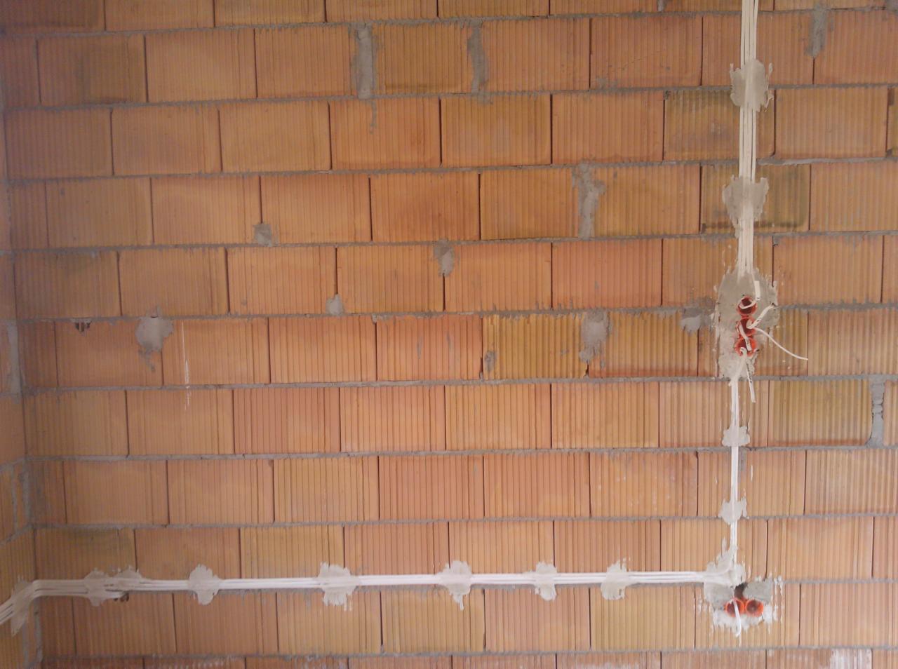 Kable elektryczne w białych rurkach, prowadzone po ścianie z czerwonej cegły, widoczne puszki z wystającymi przewodami.