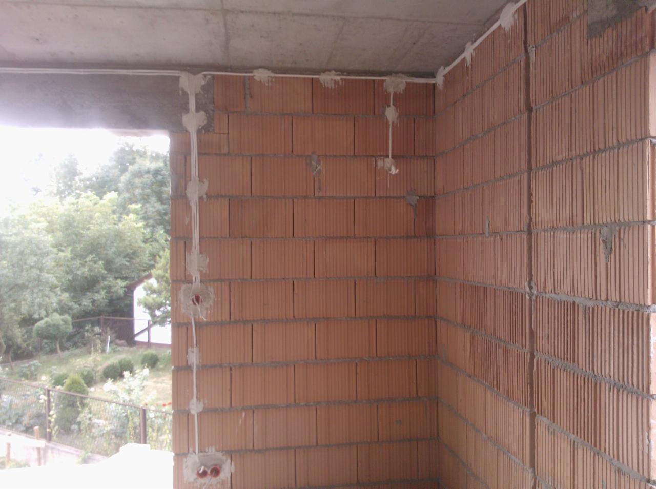 Surowa ceglana ściana z widocznymi przewodami elektrycznymi, mocowanymi gipsową zaprawą, na tle okna z widokiem na zieleń.