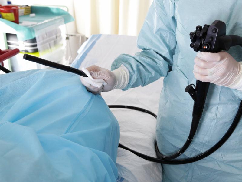Przygotowanie do kolonoskopii: lekarz w stroju ochronnym czyści końcówkę endoskopu przed badaniem pacjenta leżącego na stole zabiegowym przykrytym niebieską płachtą.
