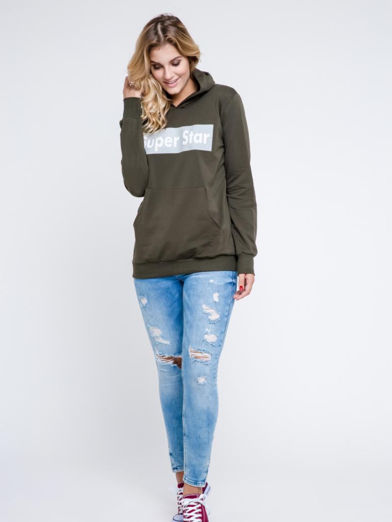 Blond modelka w oliwkowej bluzie z kapturem z napisem 'Super Star', podartych jeansach i bordowych trampkach na białym tle.
