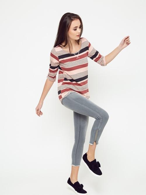 Kobieta pozuje w paski bluzce z rękawem 3/4, szarych jeansach z postrzępionymi nogawkami i czarnych wsuwanych butach z kokardką na białym tle.