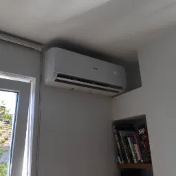 Biały klimatyzator Haier zamontowany pod sufitem w jasnym pomieszczeniu, obok okna z widokiem na drzewo i p&oacute;łki z książkami.