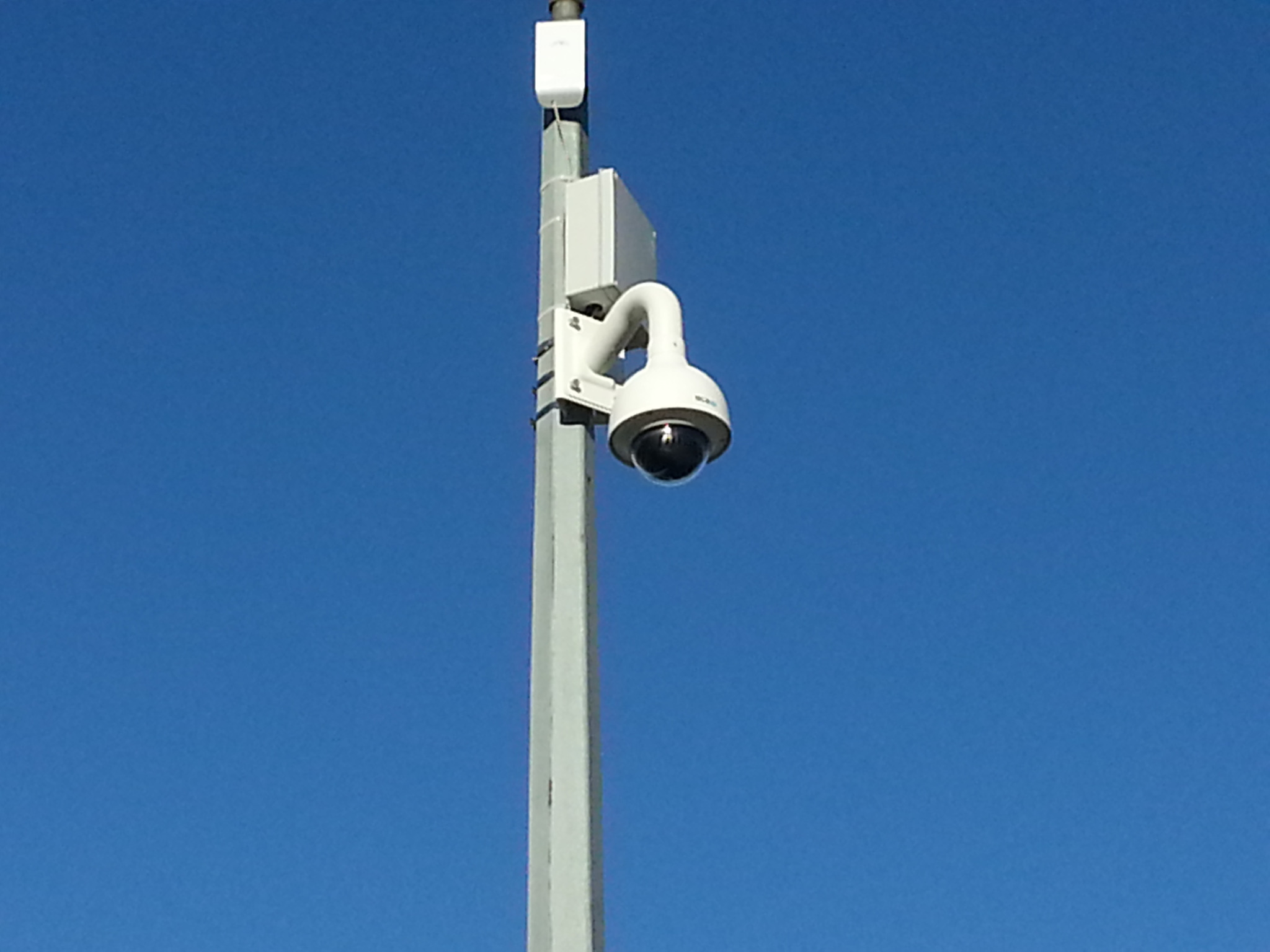 Kamera kopułkowa na szarym słupie z białymi skrzynkami montażowymi, skierowana w dół na tle błękitnego nieba.