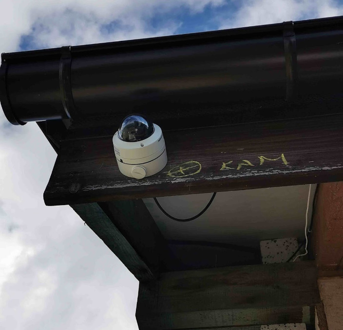 Kamera kopułkowa zamontowana pod okapem z widocznymi napisami 'KAM' wykonanymi żółtą kredą