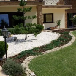 Ogród przydomowy z białym żwirem, kamieniem ozdobnym i formowanymi krzewami, oświetlony lampą ogrodową, z widocznym trawnikiem i obrzeżem z kamienia.