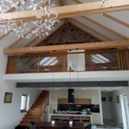 Wnętrze domu z antresolą wykończoną drewnianą balustradą, widoczne schody drewniane, kuchnia z wyspą i geometryczny żyrandol.