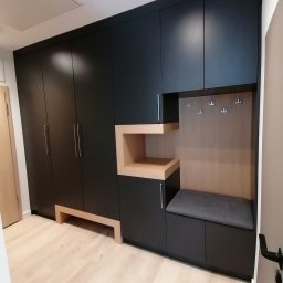 LESZEK BRZOZOWSKI - Czarna zabudowa meblowa w przedpokoju z siedziskiem i wieszakami, uzupełniona otwartą półką z jasnego drewna, na podłodze panele w odcieniach beżu.