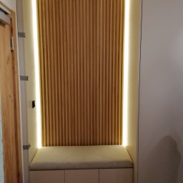 LESZEK BRZOZOWSKI - Wnęka z drewnianymi lamelami podświetlonymi taśmą LED, poniżej siedzisko z szafkami w kolorze beżowym, obok biała szafa.