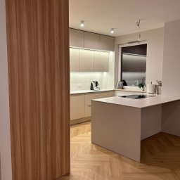 LESZEK BRZOZOWSKI - Nowoczesna kuchnia z wyspą, jasne fronty szafek, oświetlenie LED pod szafkami wiszącymi, parkiet ułożony w jodełkę, widoczna drewniana okleina na ścianie.