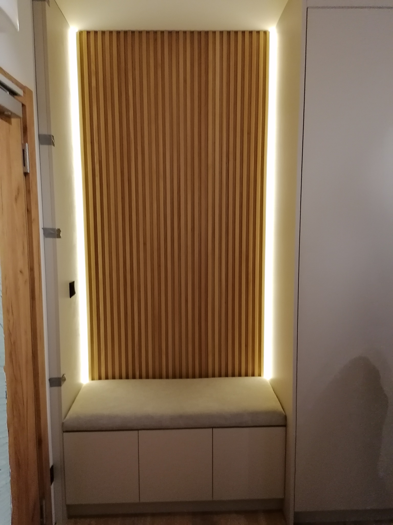 Wnęka z drewnianymi lamelami podświetlonymi taśmą LED, poniżej siedzisko z szafkami w kolorze beżowym, obok biała szafa.