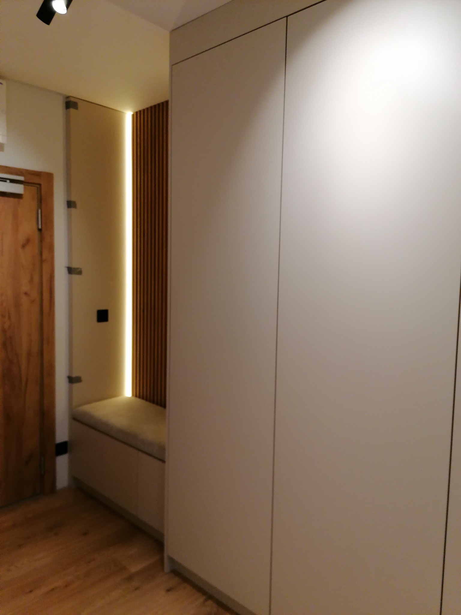 Minimalistyczna zabudowa przedpokoju w Poznaniu z dużą, jasną szafą, wnęką z drewnianymi lamelami podświetloną ledami i siedziskiem z tapicerowanym blatem.