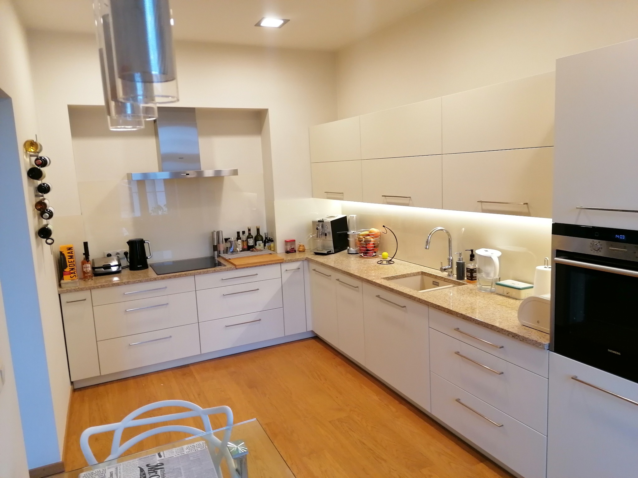 Jasna, nowoczesna kuchnia z białymi frontami i blatem imitującym kamień, oświetlona światłem LED pod szafkami wiszącymi. Widoczny fragment szklanego stołu z białym krzesłem.