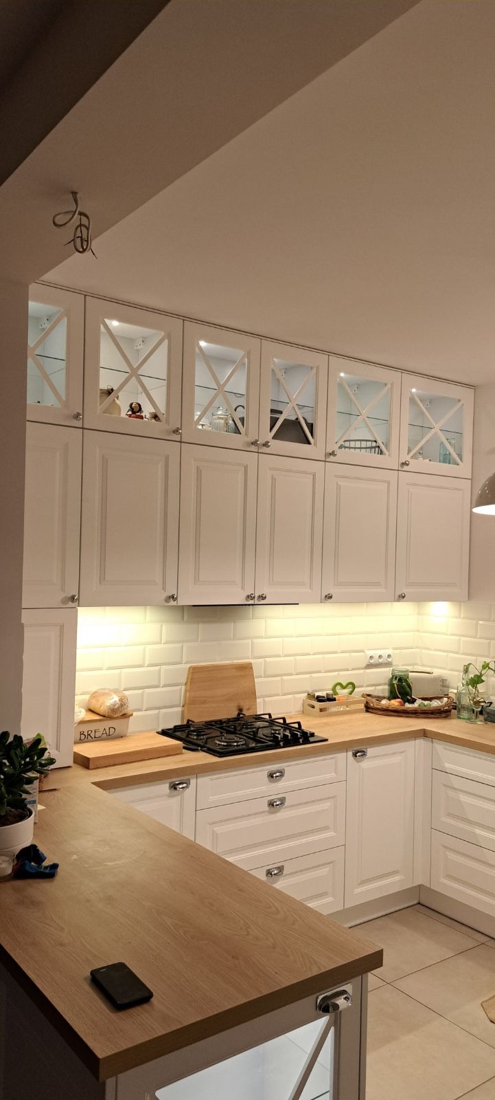 Jasna kuchnia w stylu klasycznym z drewnianym blatem, białymi szafkami z przeszkleniami w górnej części, wyeksponowany chleb w pudełku i płyta gazowa na tle białych płytek.