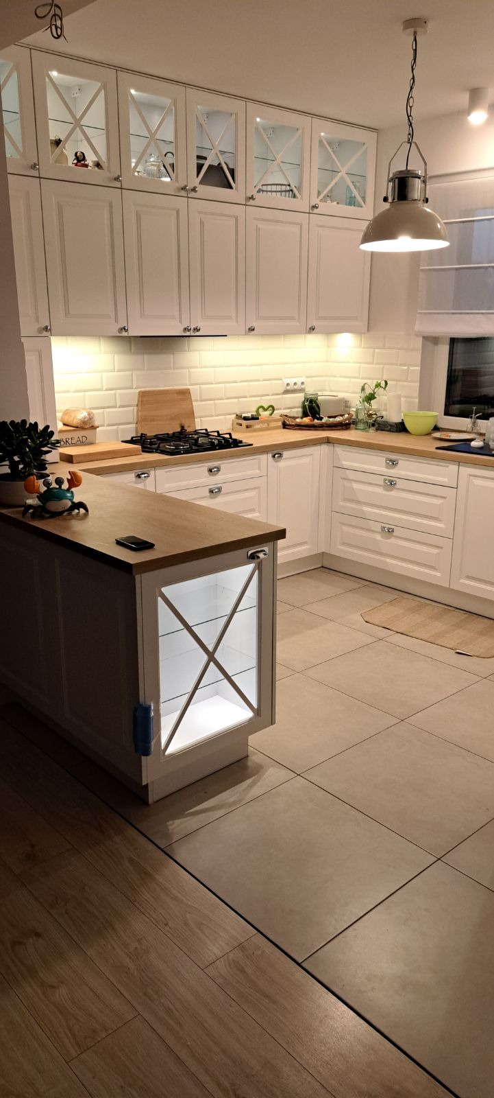 Jasna kuchnia z białymi szafkami z przeszklonymi górnymi frontami z krzyżowym szprosem, drewnianym blatem i wyspą z podświetlaną witryną, oświetlona wiszącą lampą.