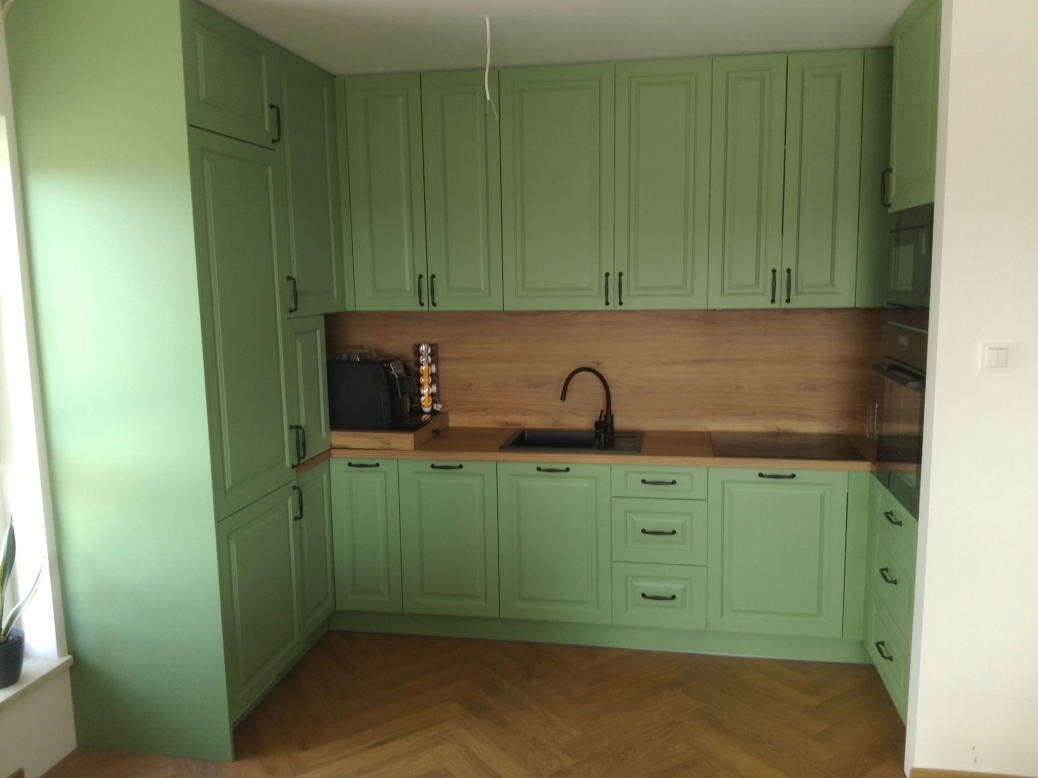 Jasnozielona zabudowa kuchenna z drewnianym blatem i parkietem w jodełkę, widok na fronty szafek i szuflad z czarnymi uchwytami.