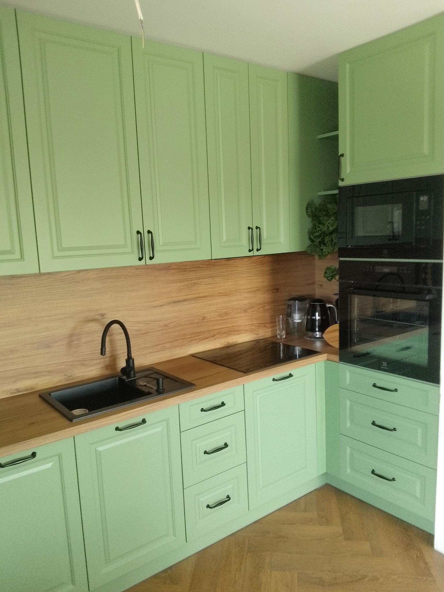 Jasnozielona kuchnia na wymiar z drewnianym blatem, czarnym zlewozmywakiem i armaturą, oraz wbudowanym piekarnikiem i mikrofalówką.