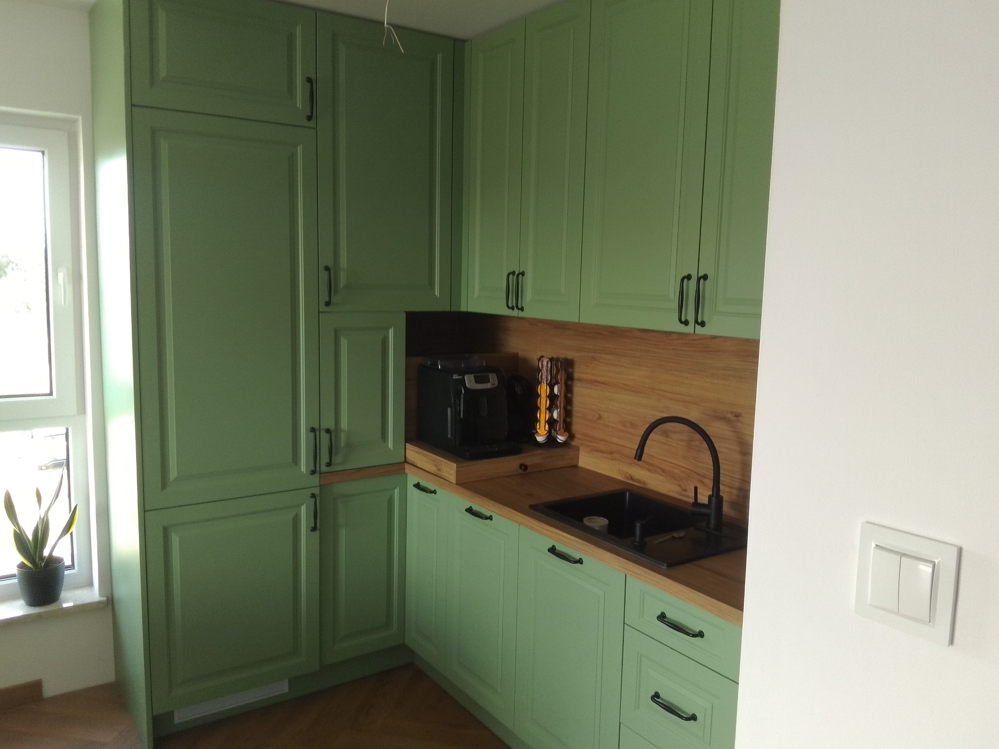 Kompaktowa kuchnia w odcieniach zieleni z drewnianym blatem i czarnym zlewem, ekspres do kawy na blacie, roślina doniczkowa na parapecie okna.