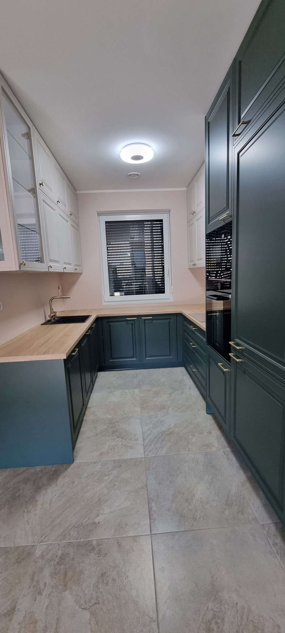 Nowoczesna kuchnia w kształcie litery U z ciemnozielonymi szafkami dolnymi i białymi górnymi, drewnianym blatem, złotą armaturą i szarymi płytkami podłogowymi, oświetlona centralnym plafonem.