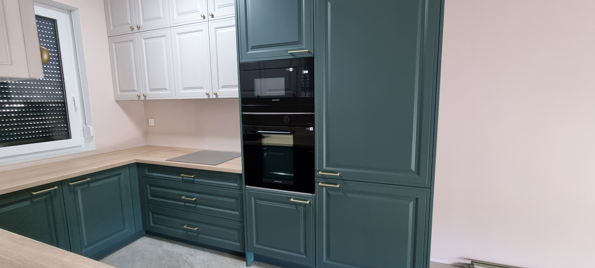 Nowoczesna kuchnia z ciemnozielonymi szafkami dolnymi i białymi górnymi, złote uchwyty, zabudowany piekarnik i mikrofalówka Samsung, jasny blat, różowe ściany.