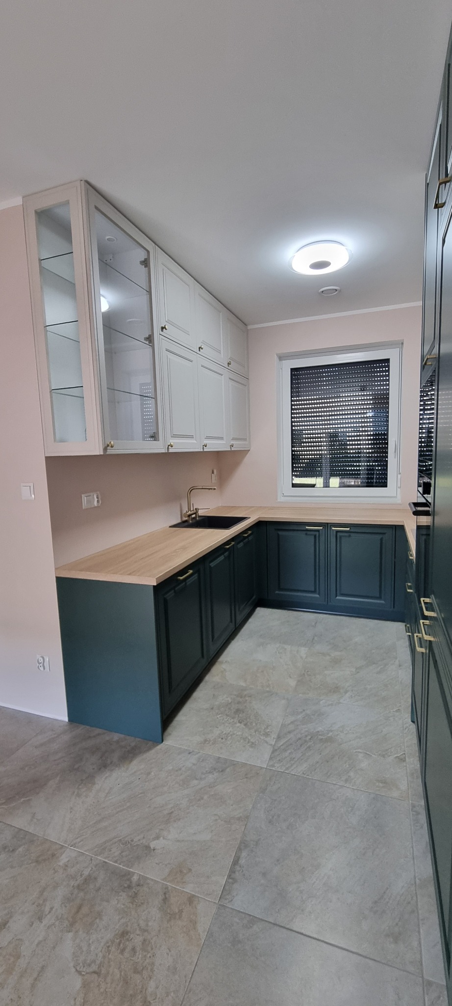 Nowoczesna kuchnia z szafkami w kolorze butelkowej zieleni i białymi szafkami wiszącymi z przeszklonymi frontami, drewnianym blatem i szarą podłogą z płytek imitujących kamień.