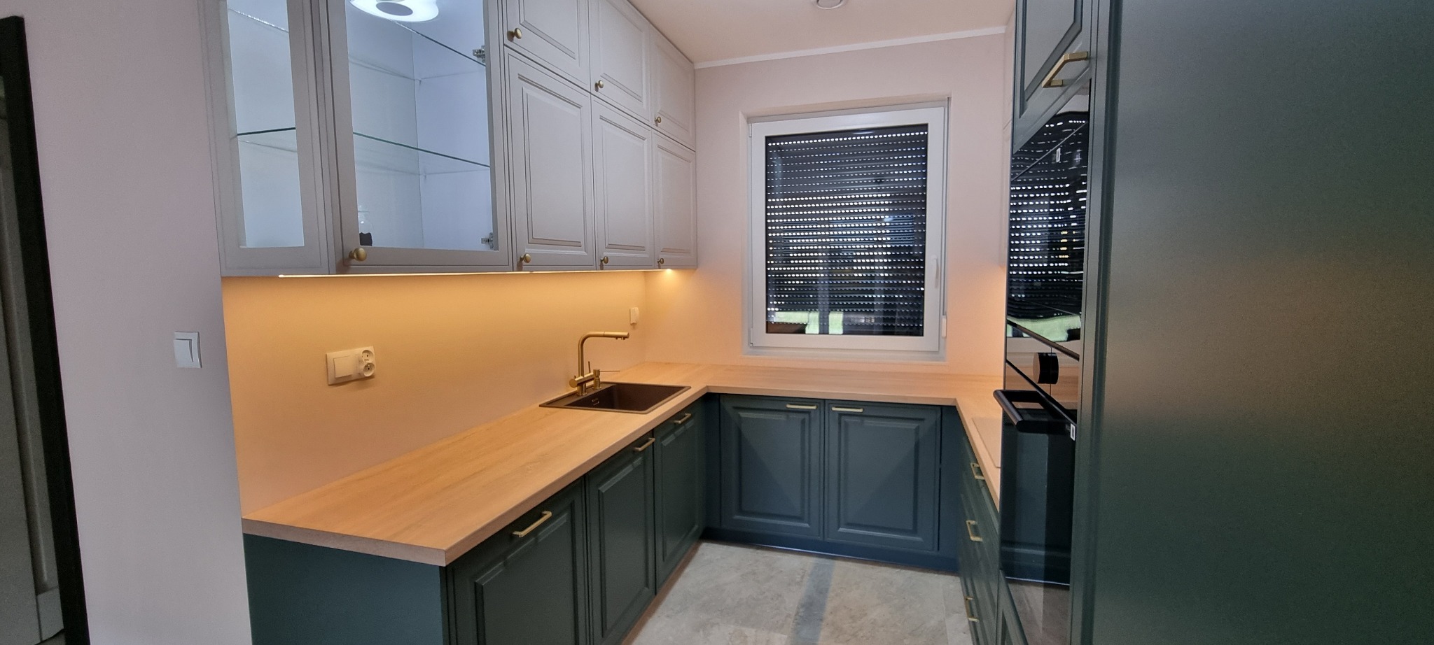 Nowoczesna kuchnia w odcieniach szarości i zieleni z drewnianym blatem, oświetleniem podszafkowym i złotą armaturą, widziana z perspektywy lekko z góry.