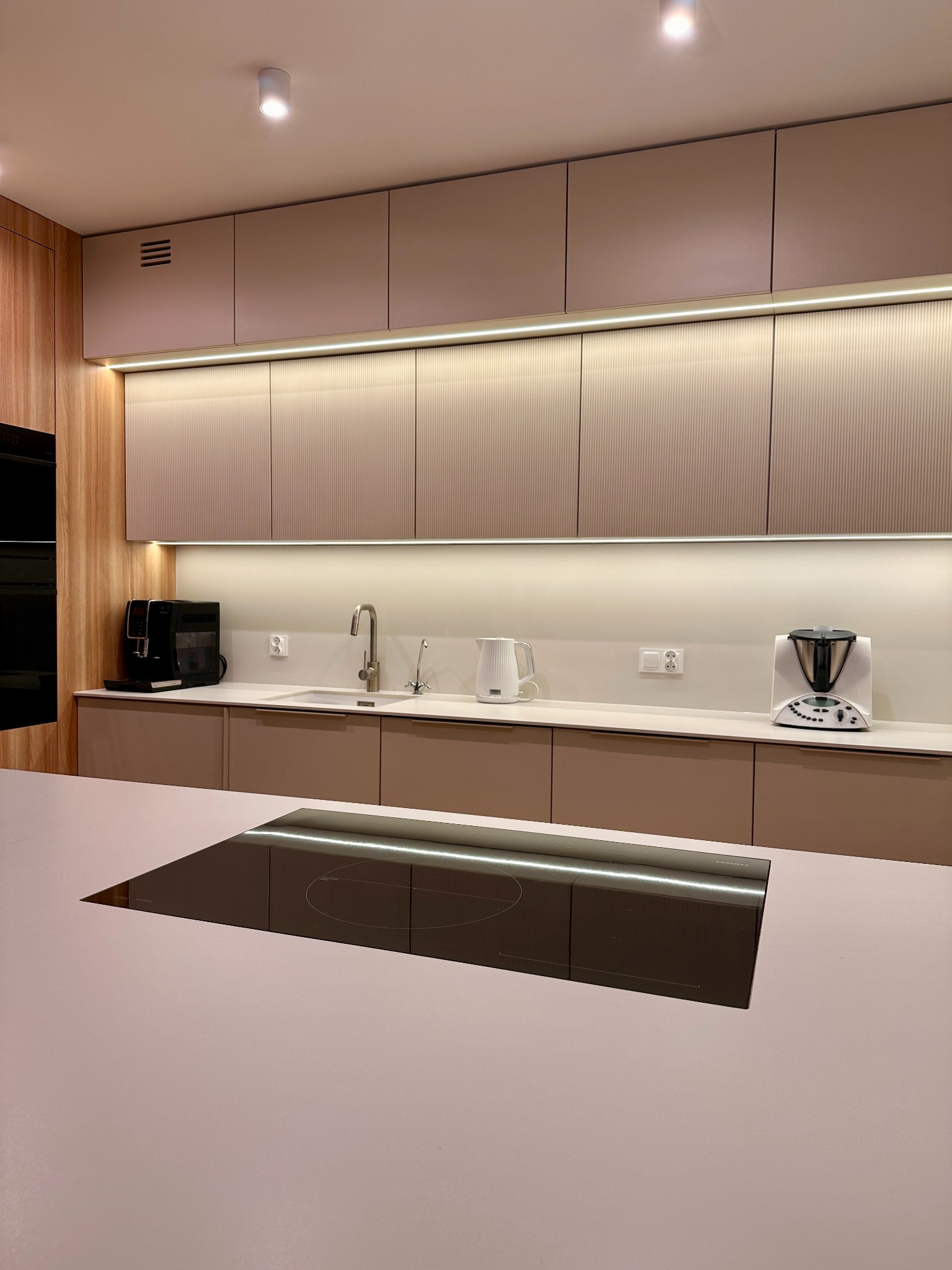 Nowoczesna kuchnia z beżowymi frontami szafek o subtelnym pionowym żłobieniu, podświetleniem LED pod szafkami wiszącymi i płytą indukcyjną wkomponowaną w jasny blat wyspy kuchennej.