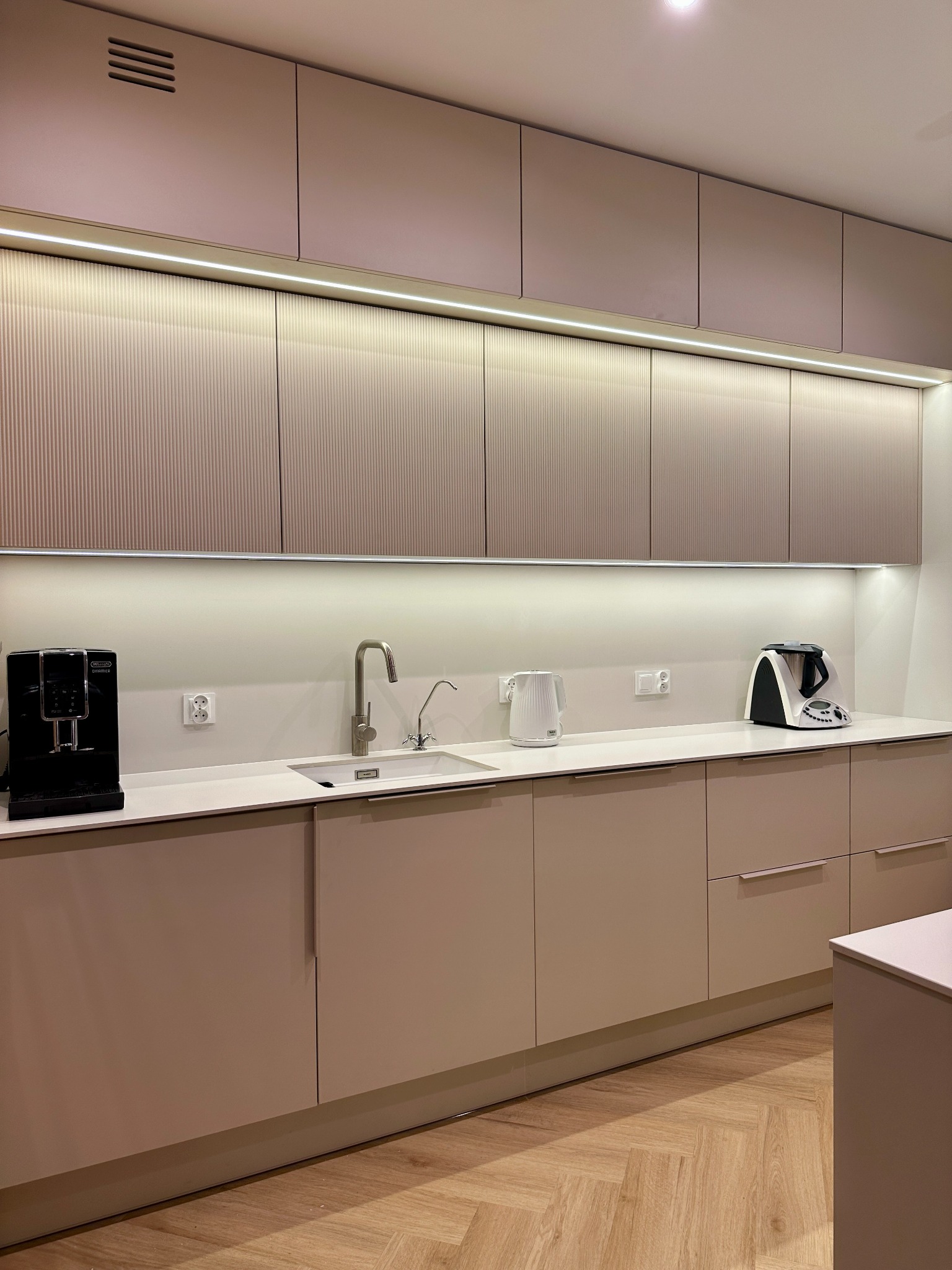 Nowoczesna kuchnia w odcieniach beżu z frezowanymi frontami szafek górnych, oświetleniem LED podszafkowym i sprzętami AGD na blacie.