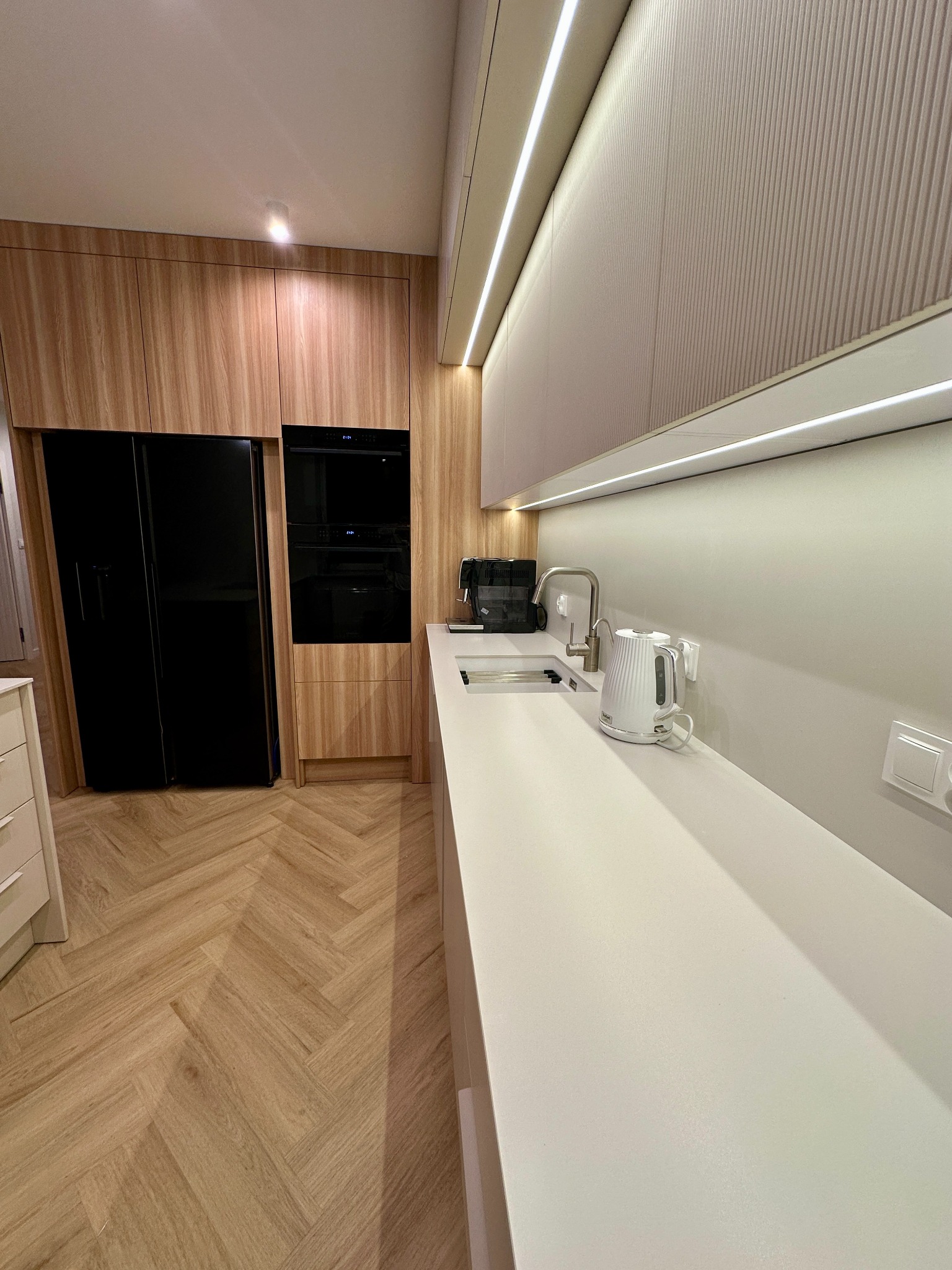 Nowoczesna kuchnia z zabudową w odcieniach drewna i bieli, czarną lodówką side-by-side i piekarnikiem w zabudowie, oświetleniem LED podszafkowym i podłogą w jodełkę.