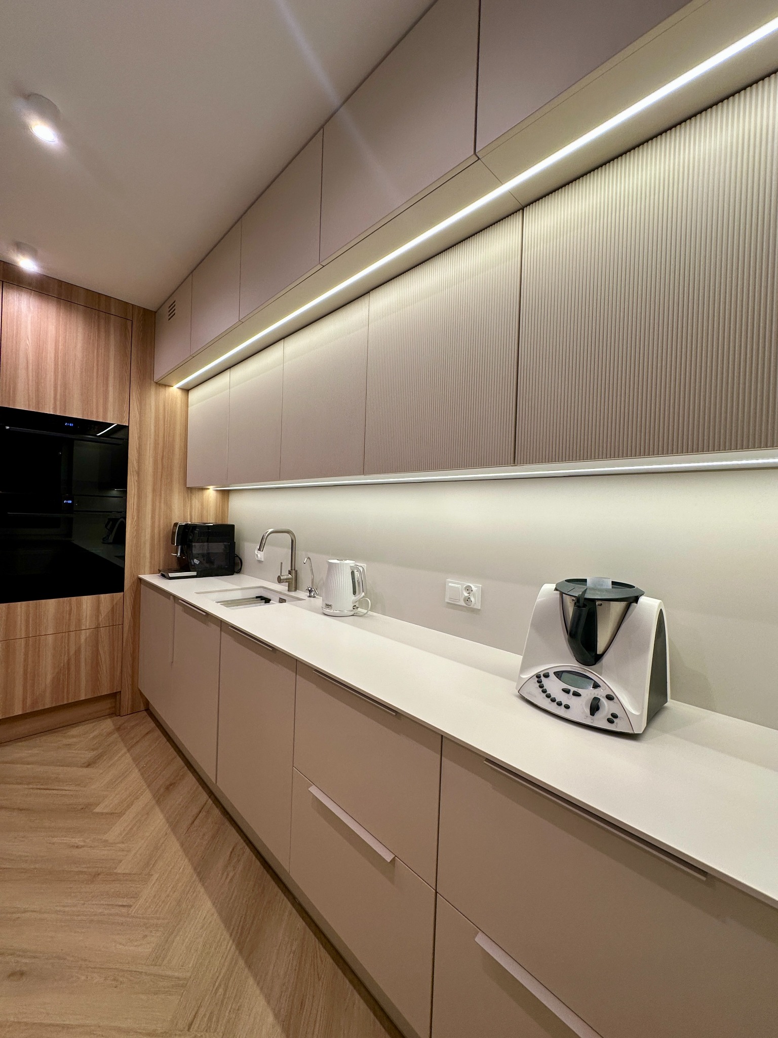 Nowoczesna, beżowo-drewniana kuchnia z oświetleniem LED w Poznaniu, z widocznymi sprzętami AGD i parkietem ułożonym w jodełkę.