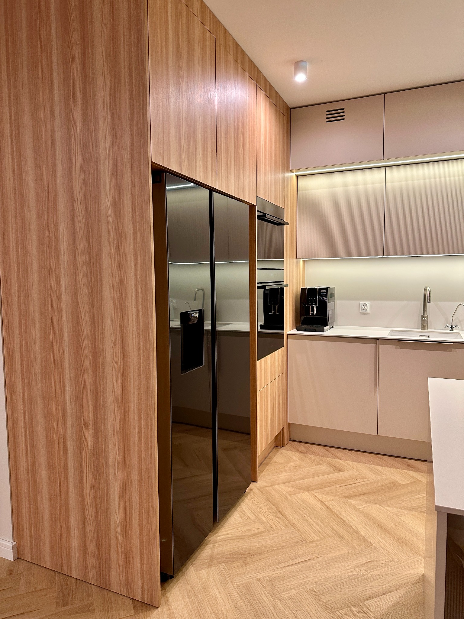 Nowoczesna kuchnia z zabudową meblową z forniru drewnopodobnego, obejmującą lodówkę side-by-side z czarnym frontem, oraz jasnymi szafkami z ryflowanymi frontami i oświetleniem LED podszafkowym.