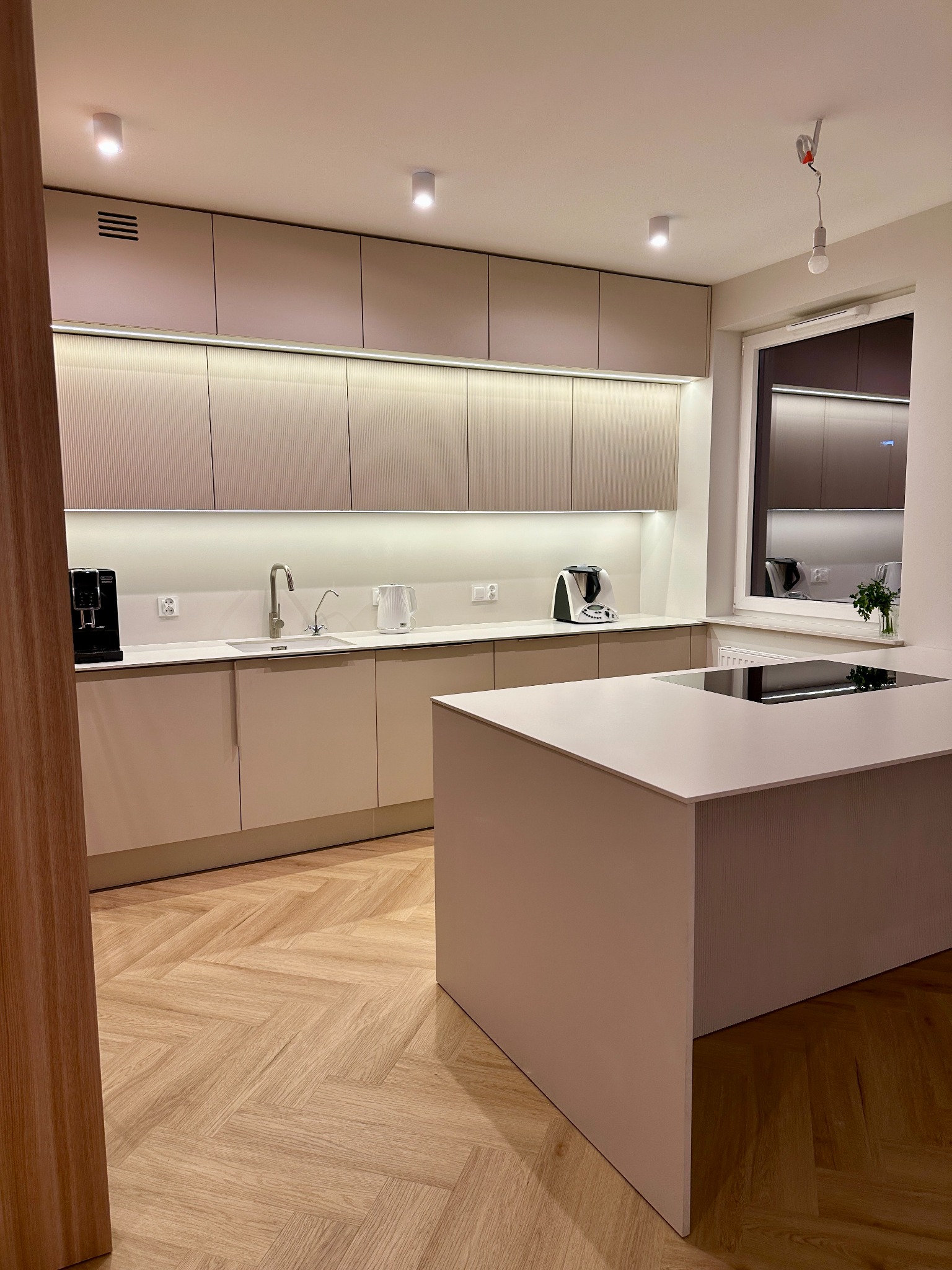 Nowoczesna kuchnia z beżowymi szafkami z ryflowanym frontem, białym blatem i wyspą, parkiet w jodełkę, oświetlenie podszafkowe i punktowe.