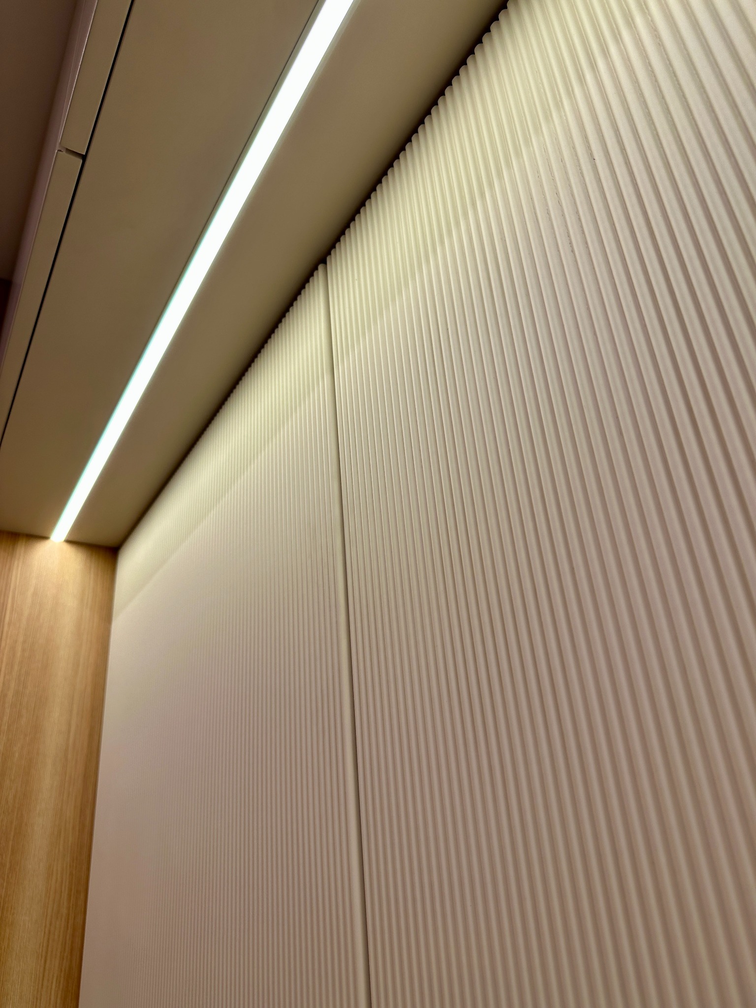 Białe, pionowo żłobkowane panele ścienne podświetlone listwą LED, z widocznym fragmentem drewnianej ściany po lewej stronie.